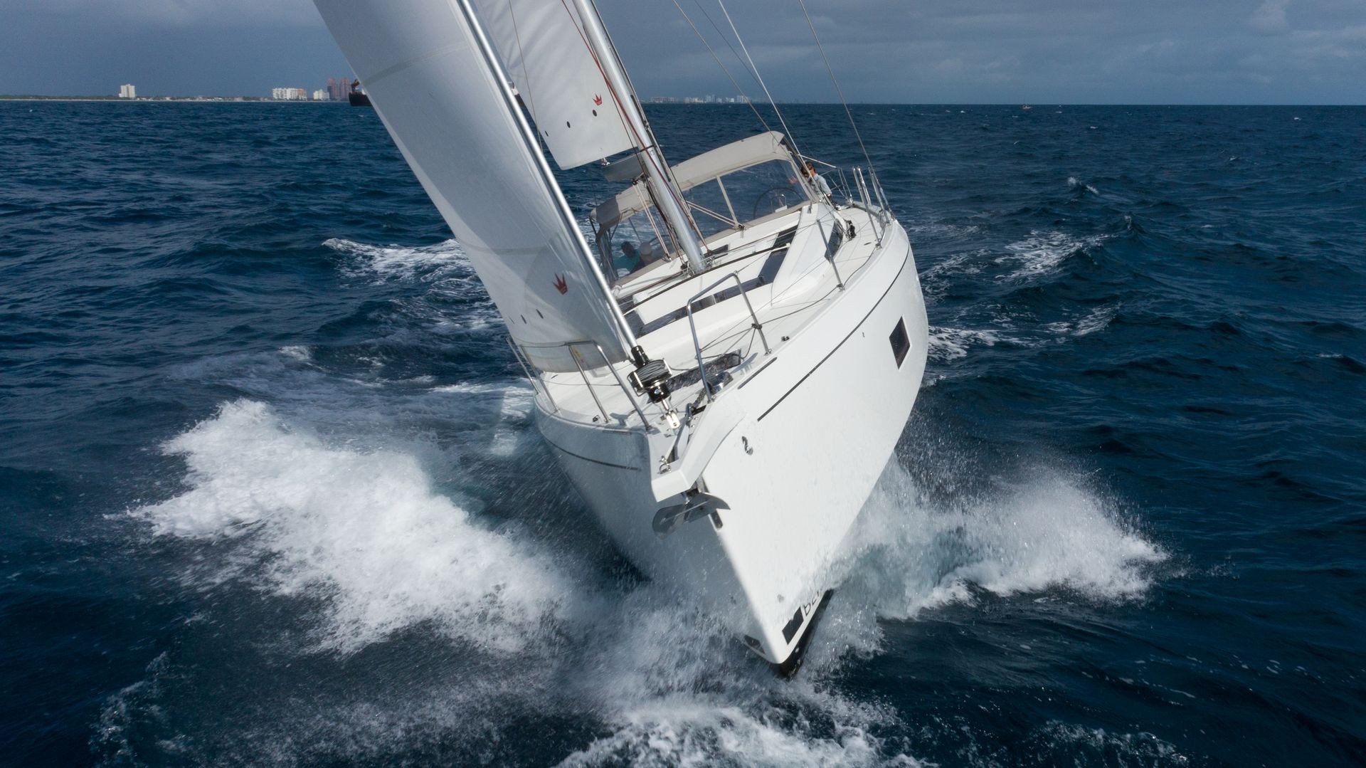 White sailboat cutting through the waves in open sea with city skyline visible in the distant background.